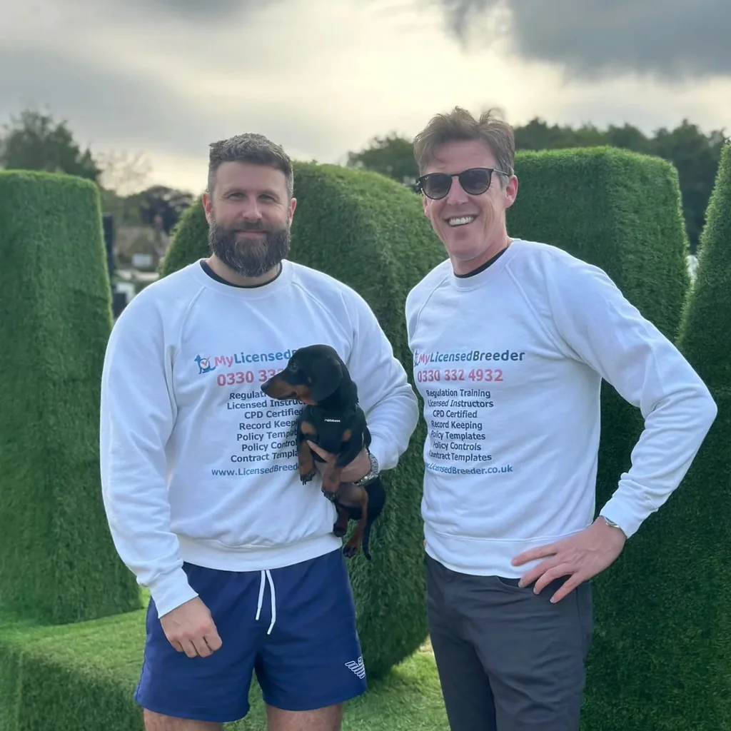 Two people wearing My Licensed Breeder tops outdoors, with a dog held between them